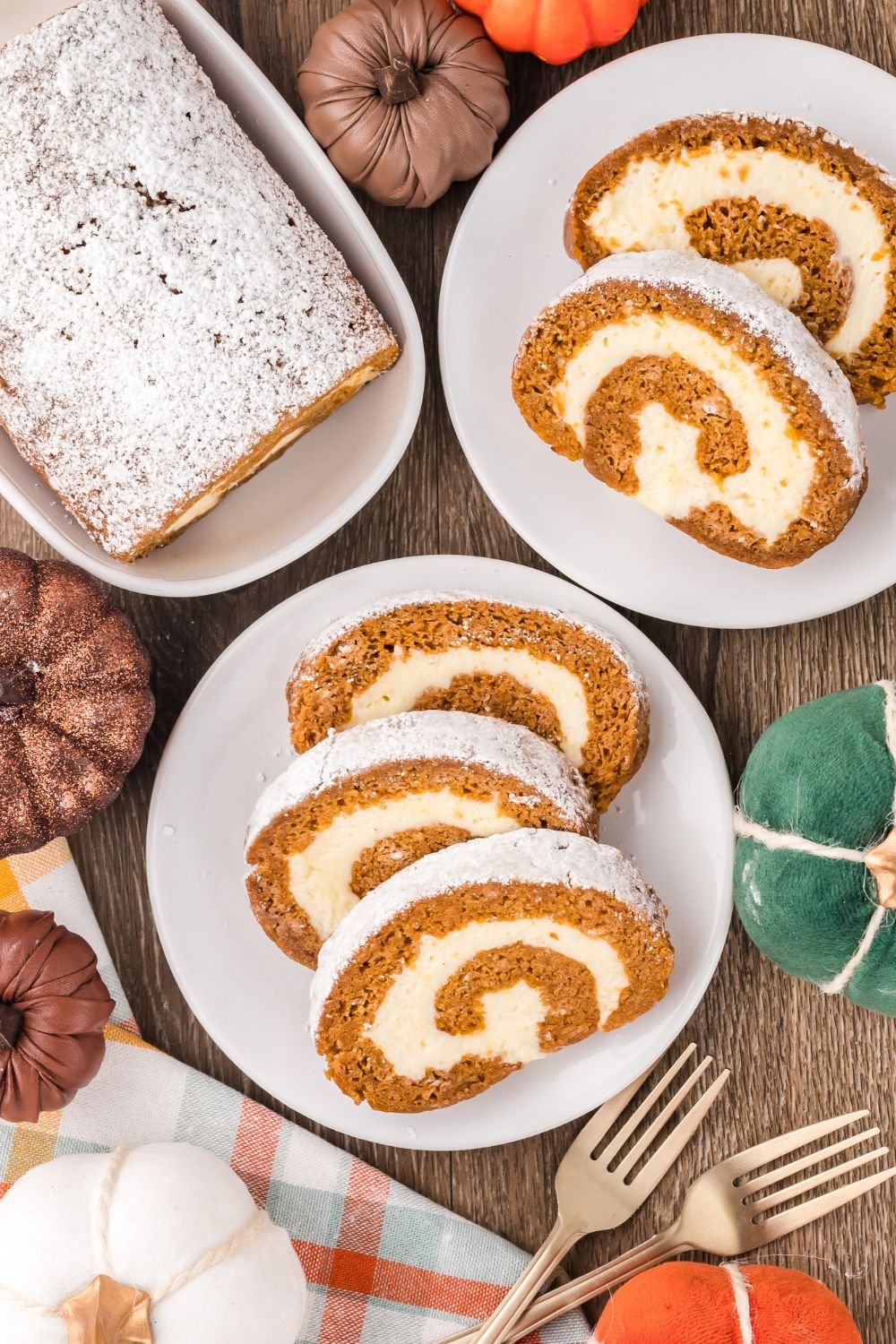 fall rustic wooden table with faux pumpkins and plates with slices of Pumpkin Cake Roll on it, and a platter to the back with more of the pumpkin cake on it 