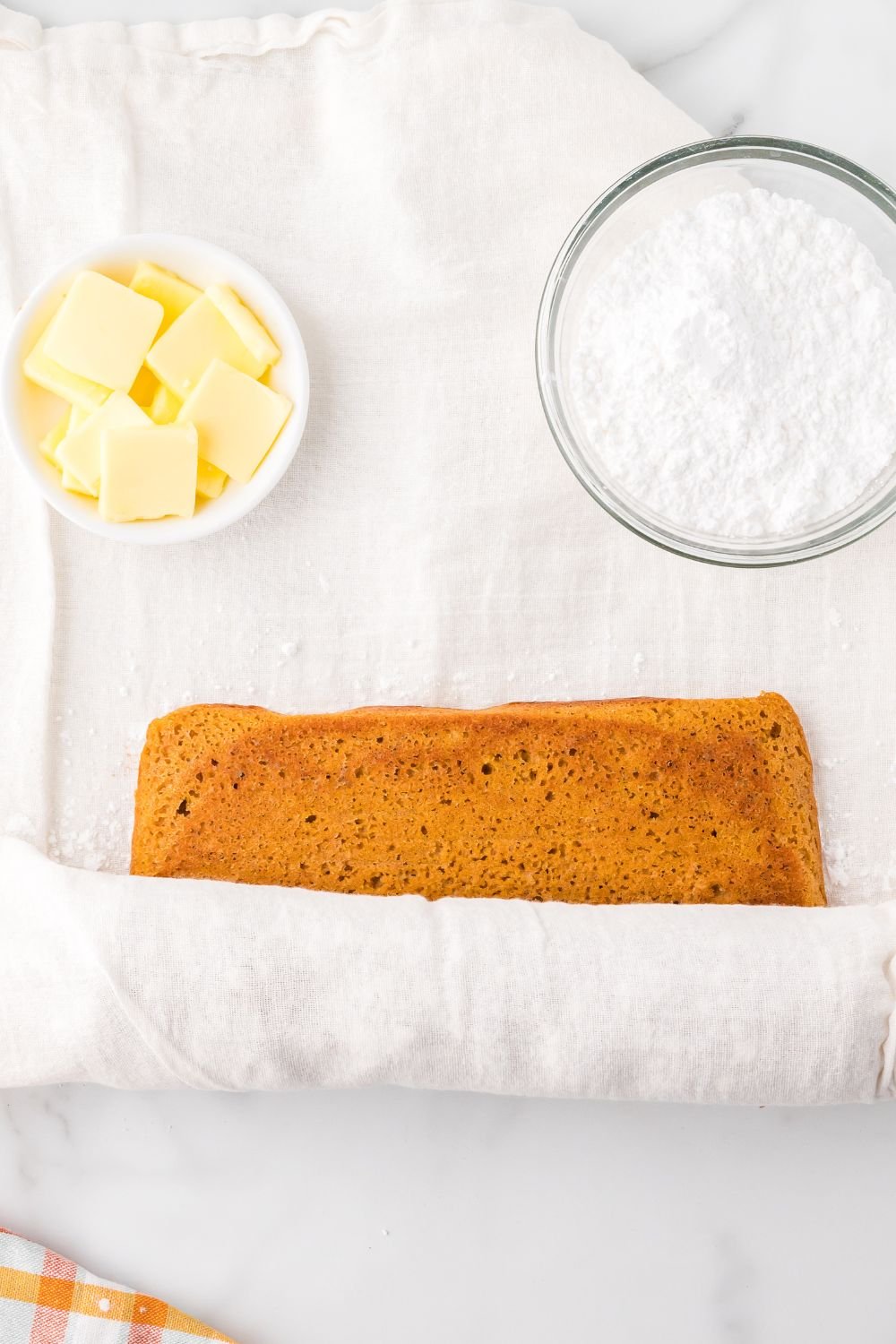 rolling up a Pumpkin Cake Roll with parchment paper. To the side are two small bowls one with sliced butter the other with powdered sugar