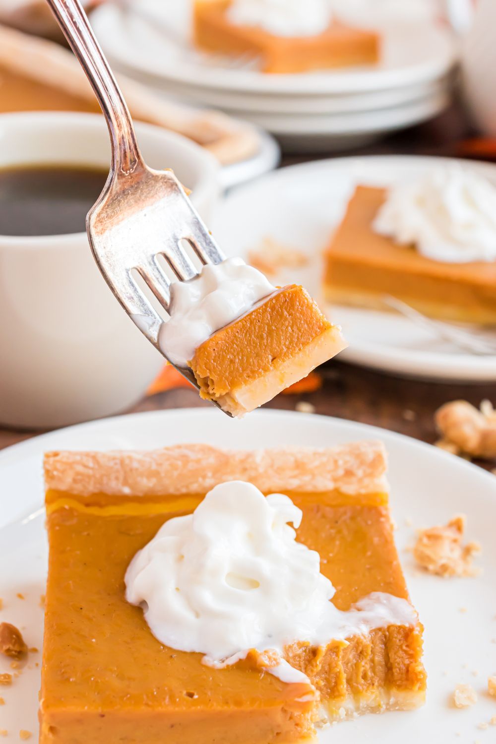 a handful of plates spread out on a wooden table with pumpkin pie topped with whipped cream and coffee cup in the center of table. Then one slice of pie has a fork with some of the pie and whipped cream on the fork. 