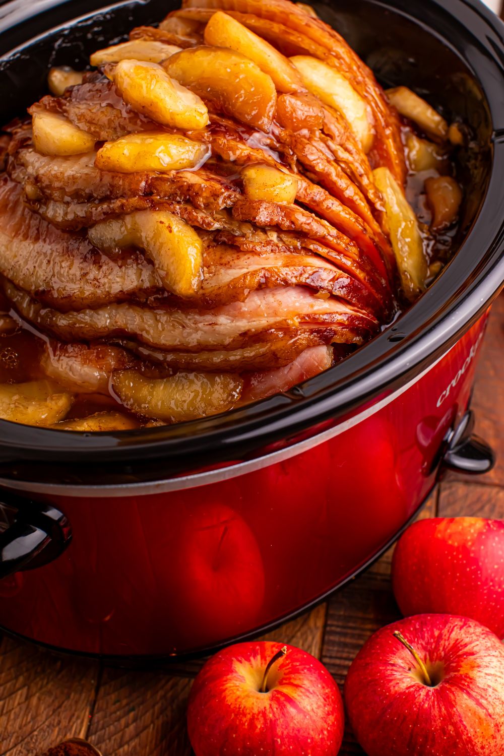 red slow cooker on a wooden table with a spiral ham with apples on top, and whole apples in front of table