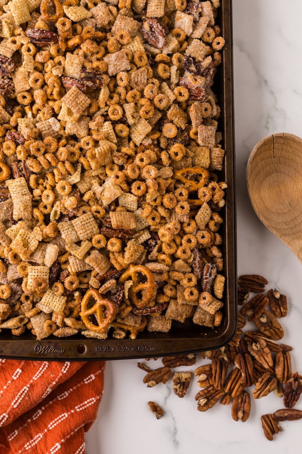looking down at counter with a sheet pan full of Pumpkin Spice Chex Mix and some nuts scattered on counter with a wooden spoon to the side