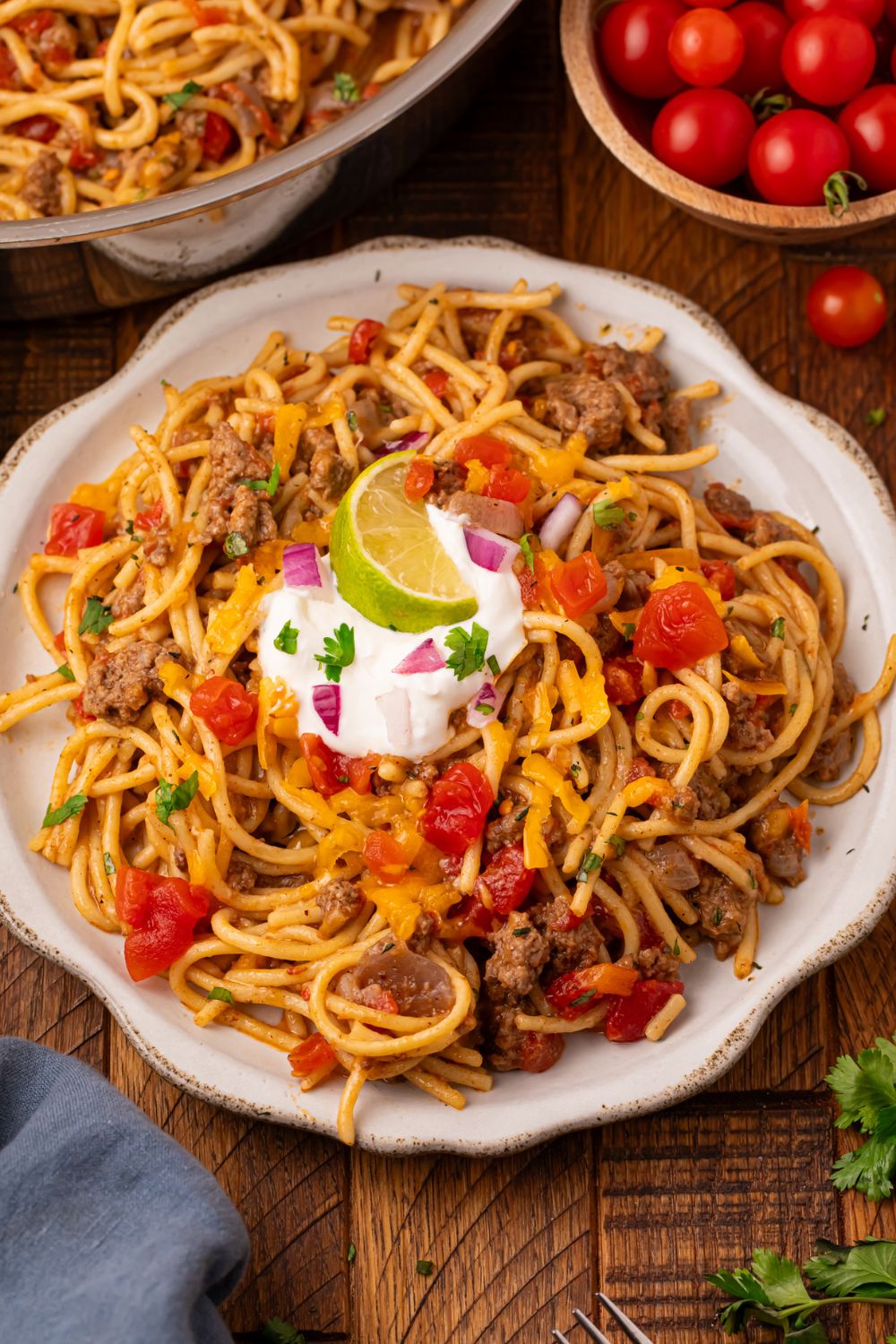 Table set up for serving up dinner with a bowl of tomatoes to side, skillet with more pasta, and a large white place and topped with sour cream, and a lime wedge 