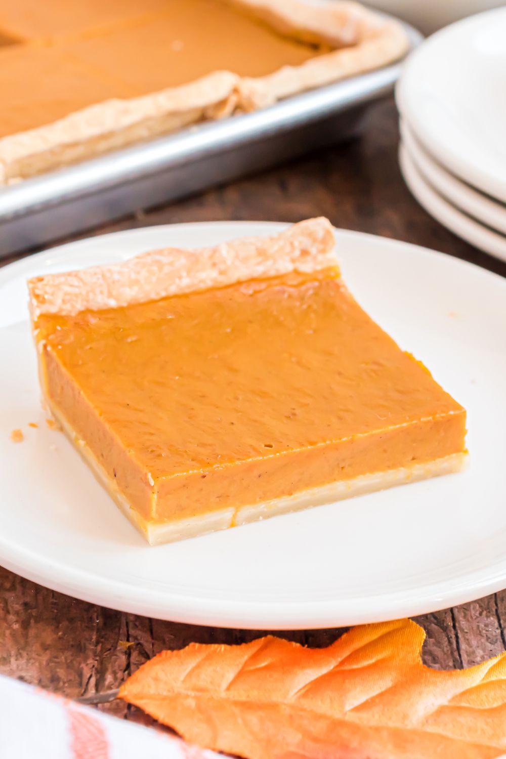 slice of Sheet Pan Pumpkin Pie on a white plate with a stack of white plates to side on wooden table and sheet pan pie behind it on table 