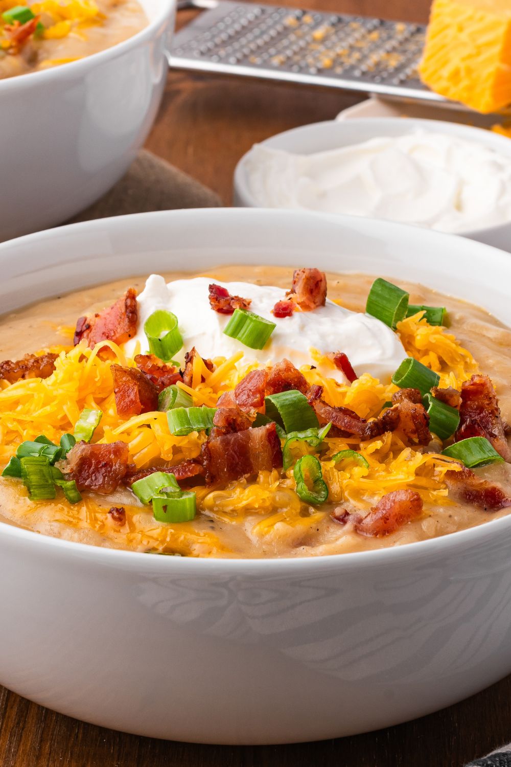 side view of table with two stacks of white bowls full of Crockpot Loaded Baked Potato Soup topped with shredded cheese, green onions and back bits and sour cream. Behind the table is a bowl of sour cream 