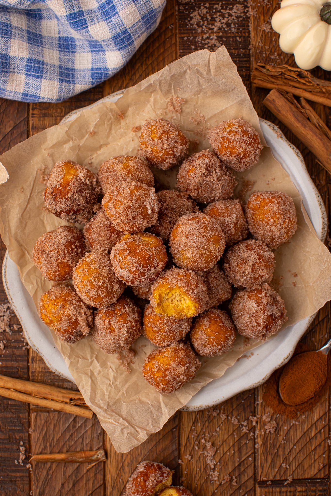 Up close overhead shot oif the Pumpkin Pie Donut Holes scattered on a parchment lined white platter on a wooden table. The top donut hole has a bite taken out of it. 