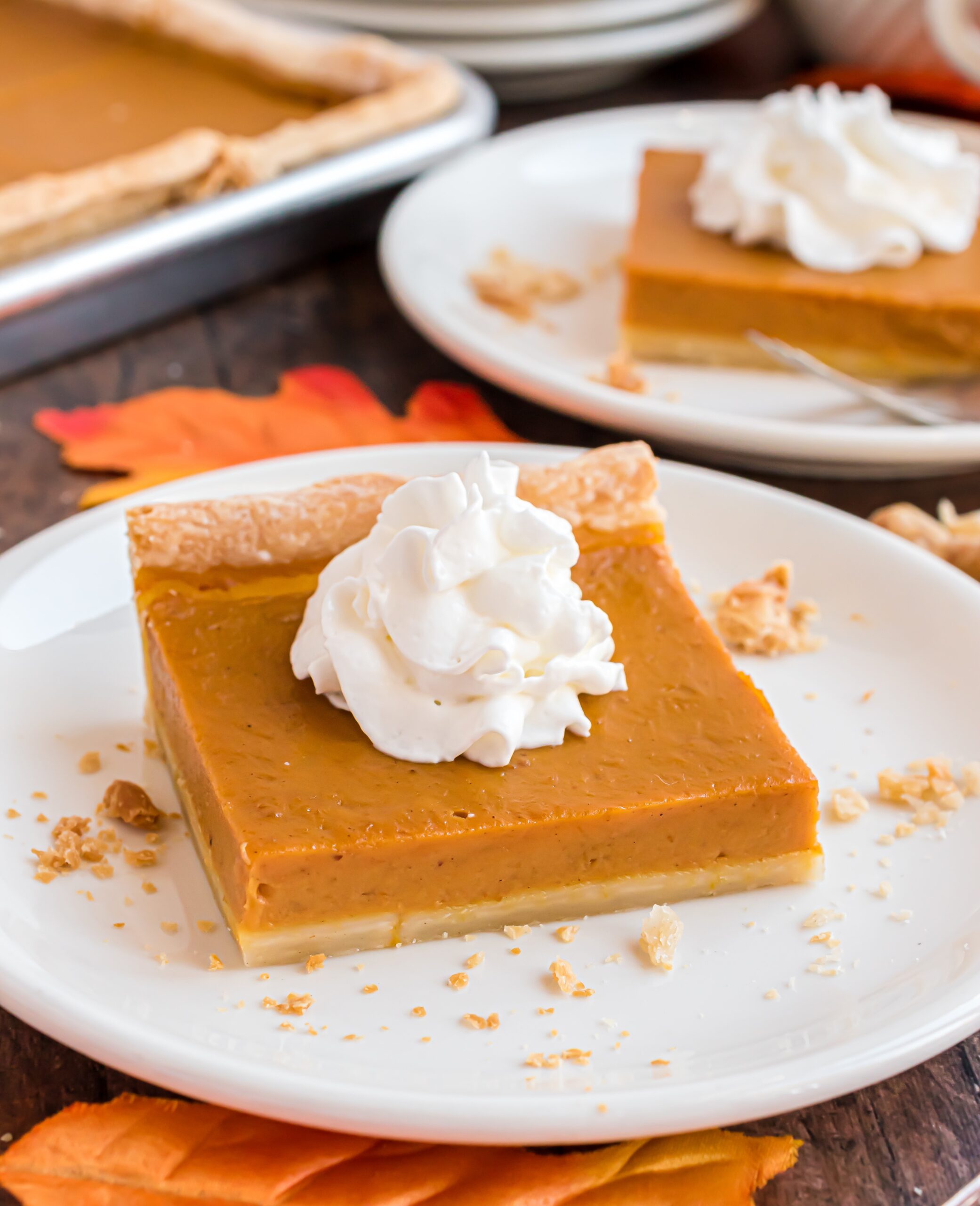 wooden table decorated for the fall season with white plates topped with slices of pumpkin pie topped with whipped cream.