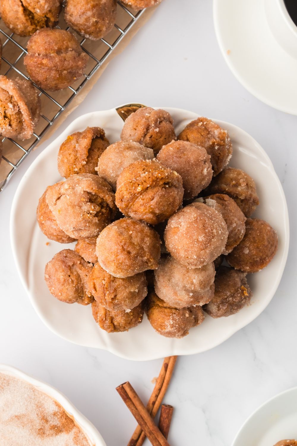 Pumpkin Spice Donut Holes on a white plate sitting on table with cinnamon sticks to side, and more donuts on tray in background 