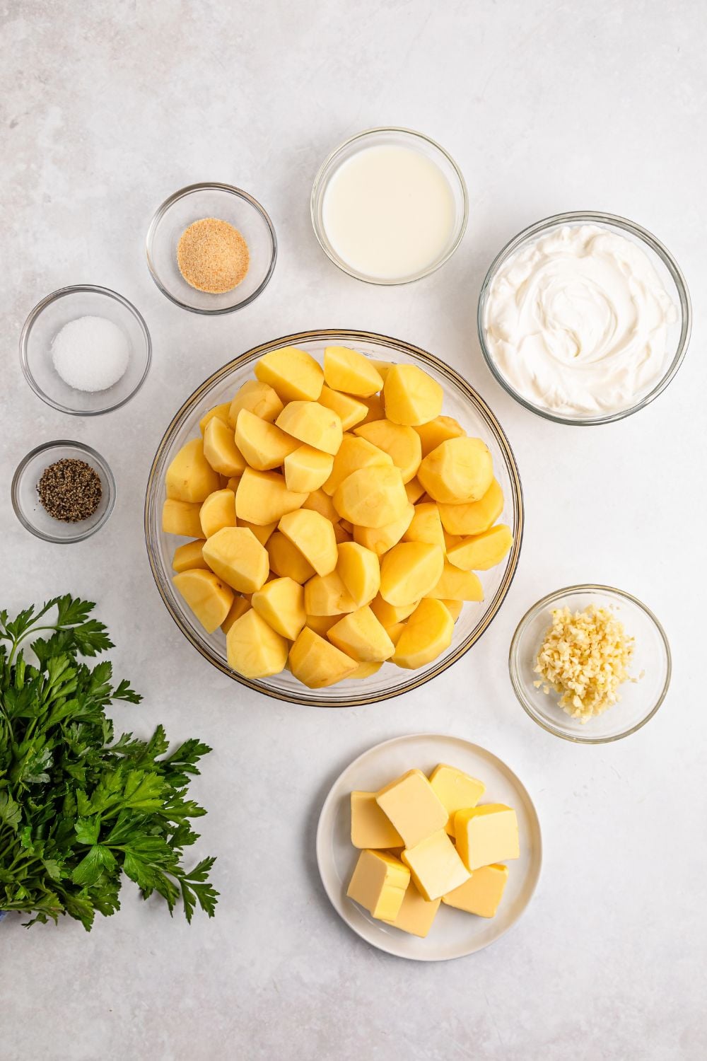 all the ingredients for Garlic Mashed Potatoes on a white table in small single serve bowls and a bundle of parsley to the side