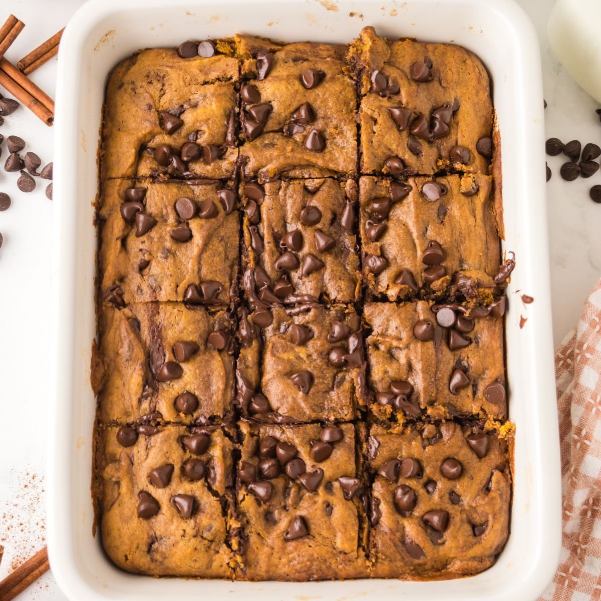 Brown Butter Pumpkin Chocolate Chip Bars in a ceramic casserole dish on table with cinnamon sticks and chocolate chips around table 