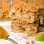Caramel Apple Pie Bars stacked on top of one another with crumble around white table and a slice of green apple to side