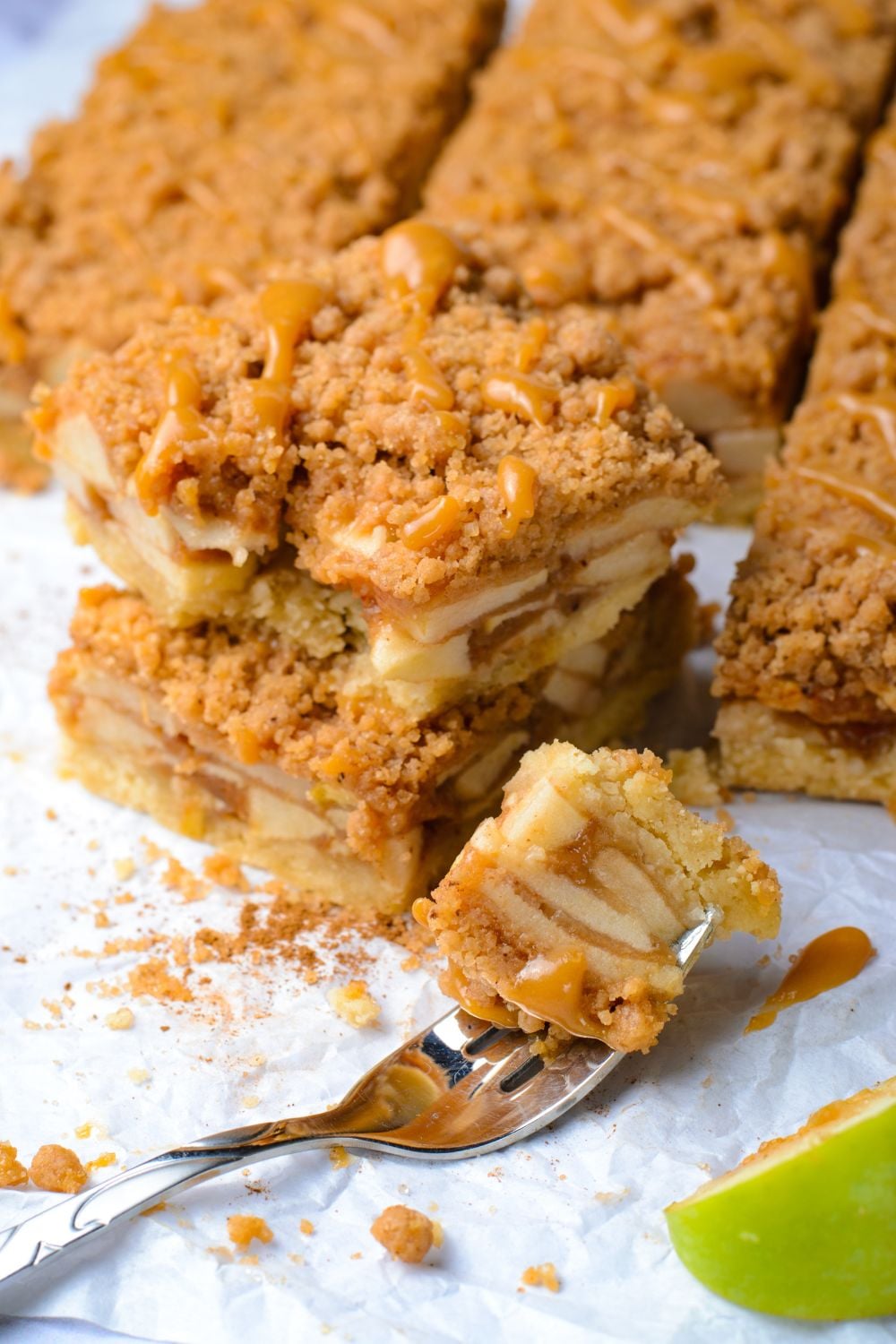 Caramel Apple Pie Bars spread out on a table with a bite taken out of the bar on a fork to side on table with a slice of green apple