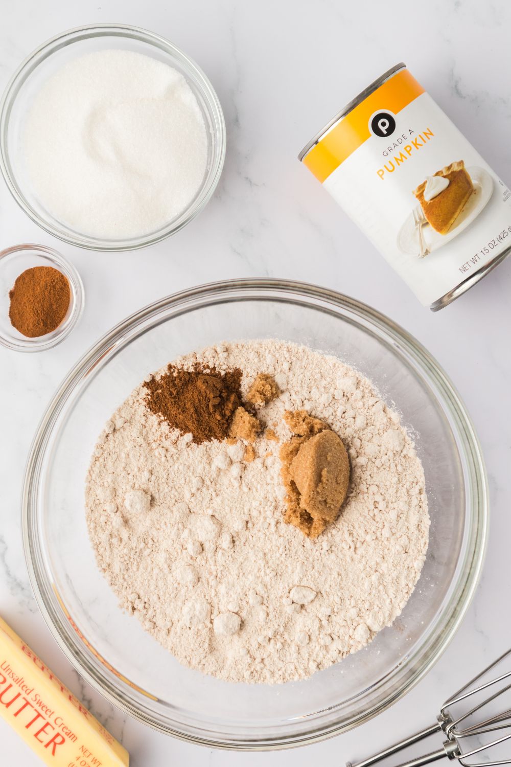 dry ingredients for making pumpkin donuts in a glass bowl with a can of pumpkin puree to the side and a stick of butter