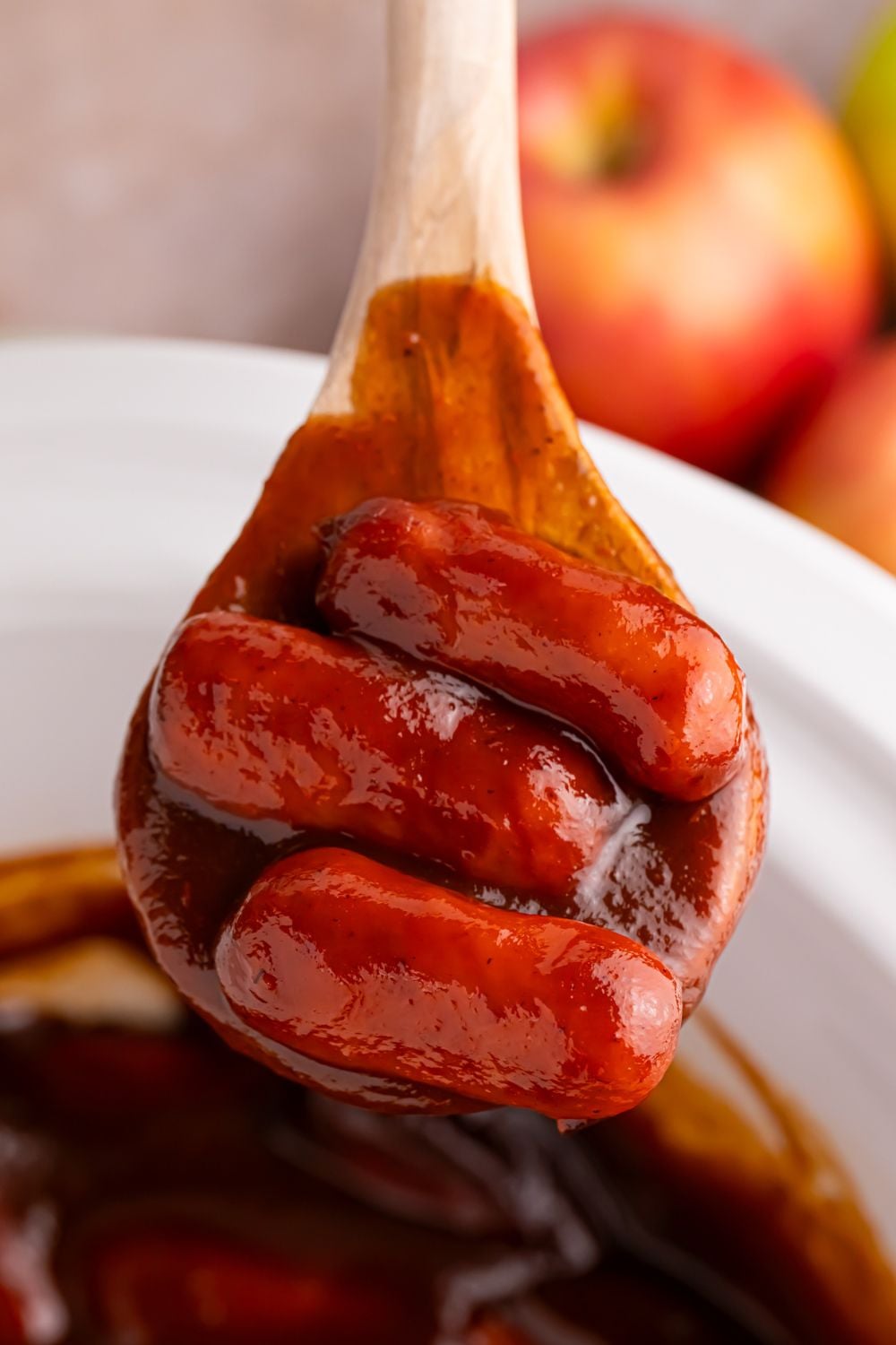 wooden spoon with some Crockpot Apple Butter BBQ Little Smokies on it with apples behind it on the table 