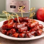 Crockpot Apple Butter BBQ Little Smokies on a white plate with toothpicks in it, with a slow cooker behind it on the table