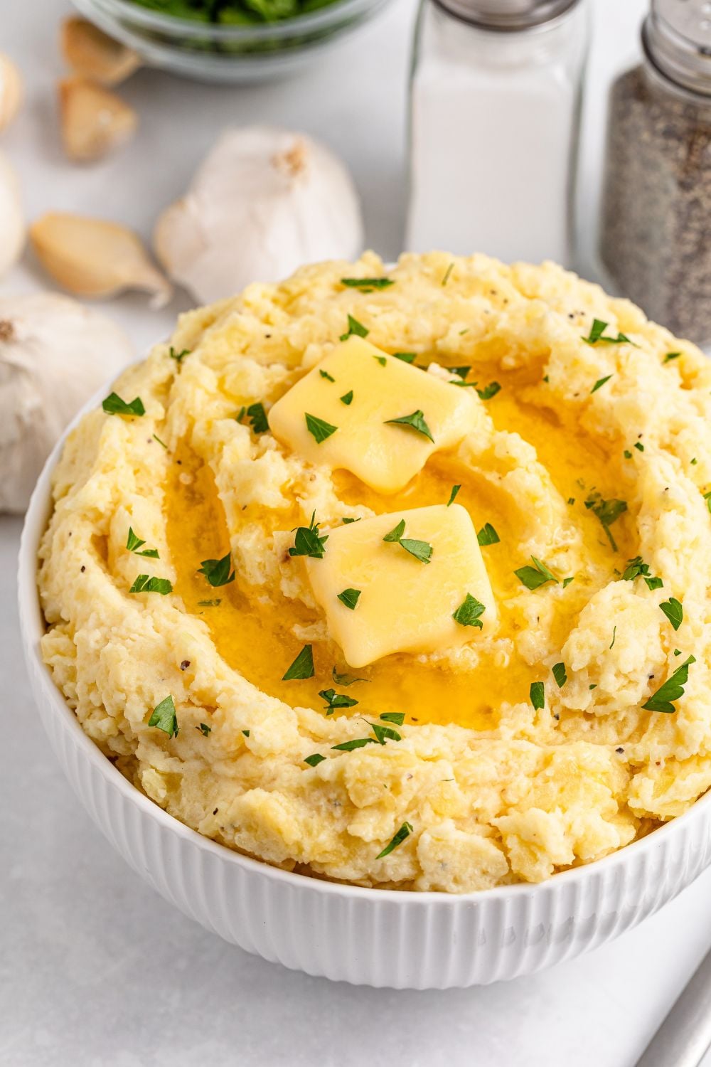 White table with salt and pepper shakers, a handful of garlic cloves, and a white bowl full of Garlic Mashed Potatoes