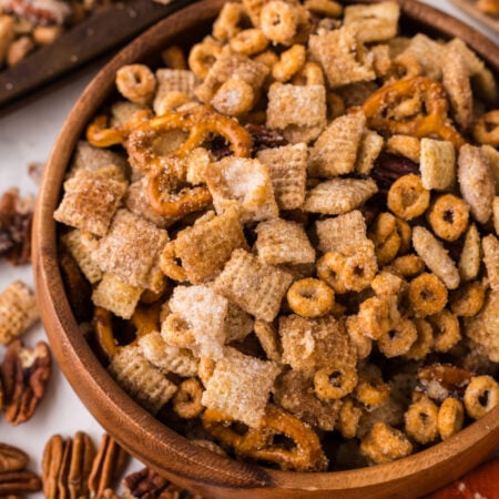 Pumpkin Spice Chex Mix in a wooden bowl on table with more mix scattered on table with a towel to the side of the bowl