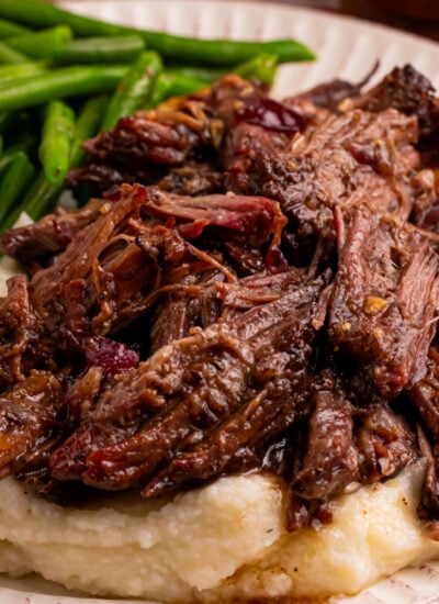 Cranberry Pot Roast shredded on a white plate with mashed potatoes under and a side of green beans