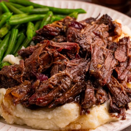 Cranberry Pot Roast shredded on a white plate with mashed potatoes under and a side of green beans