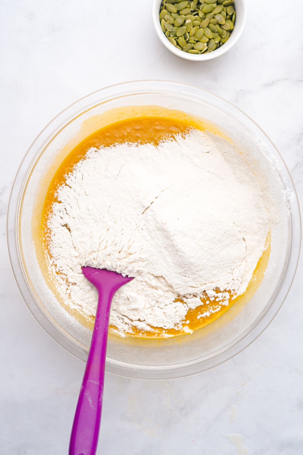 pumpkin bread batter in a glass bowl on table with flour on top and a purple spatula 