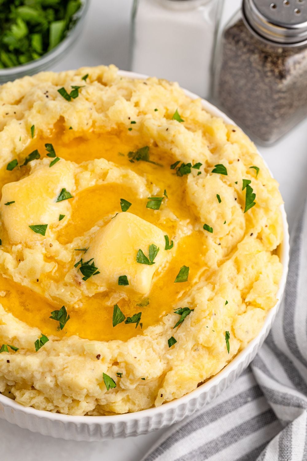 view of part of a white bowl on table full of Garlic Mashed Potatoes with melted butter and chopped parsley on top. A salt and pepper shaker are to the side of the bowl 