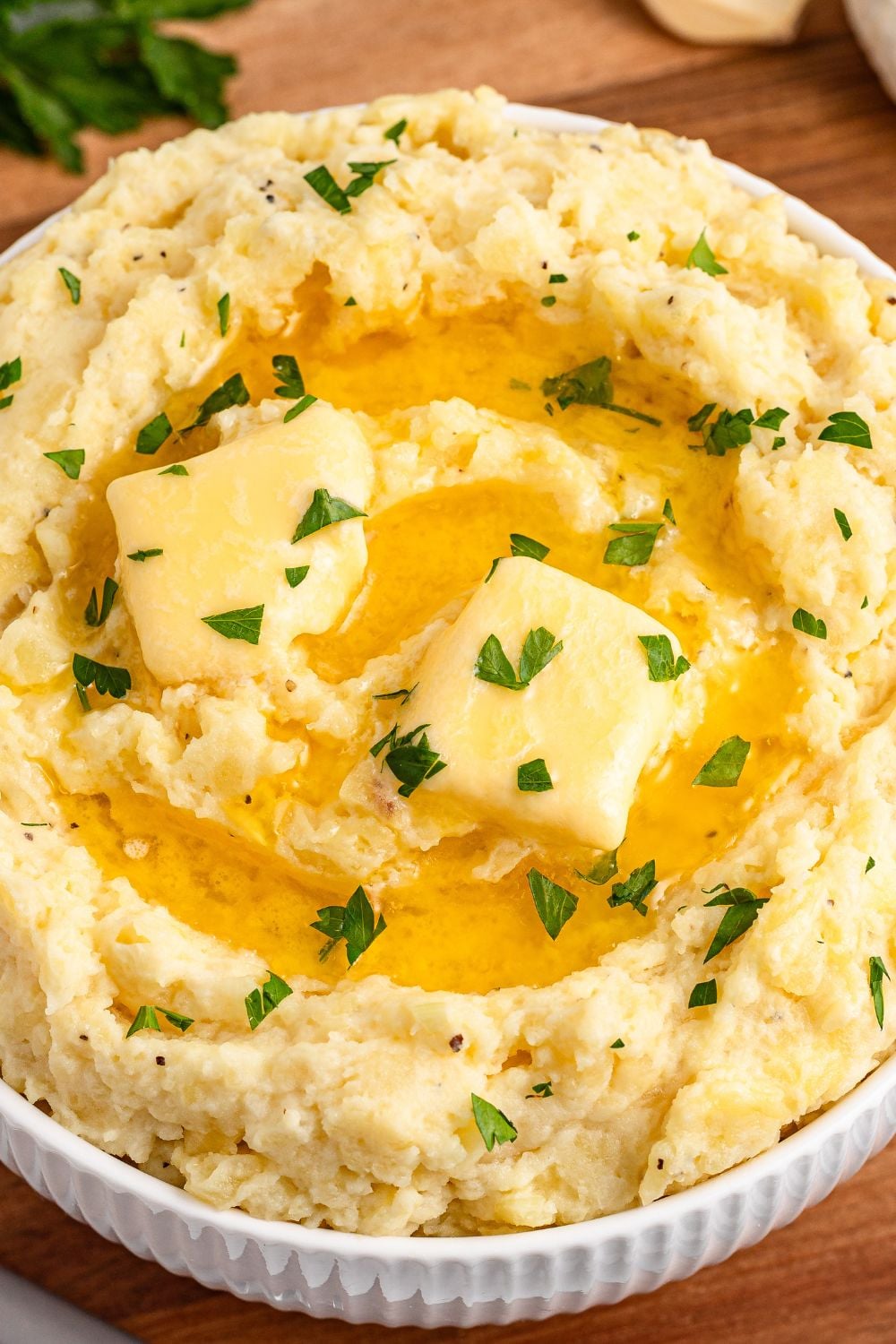 close up look of Garlic Mashed Potatoes in a white bowl on wooden cutting board. The potatoes are topped with melted butter and fresh parsley