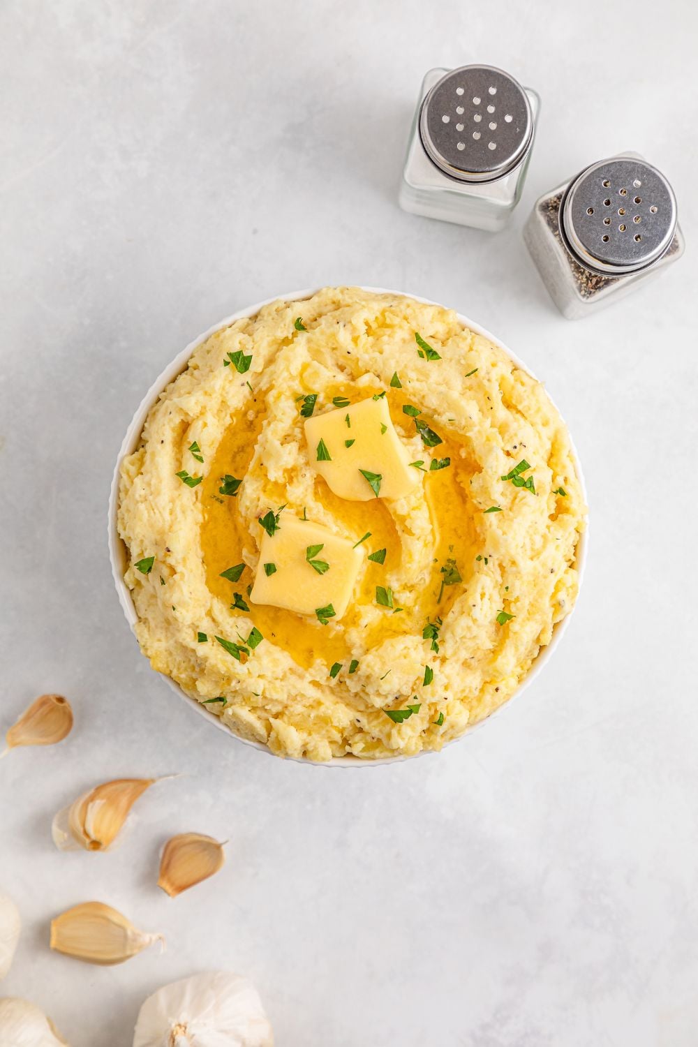 large white bowl on table full of Garlic Mashed Potatoes and garlic to side and salt and pepper shakers 