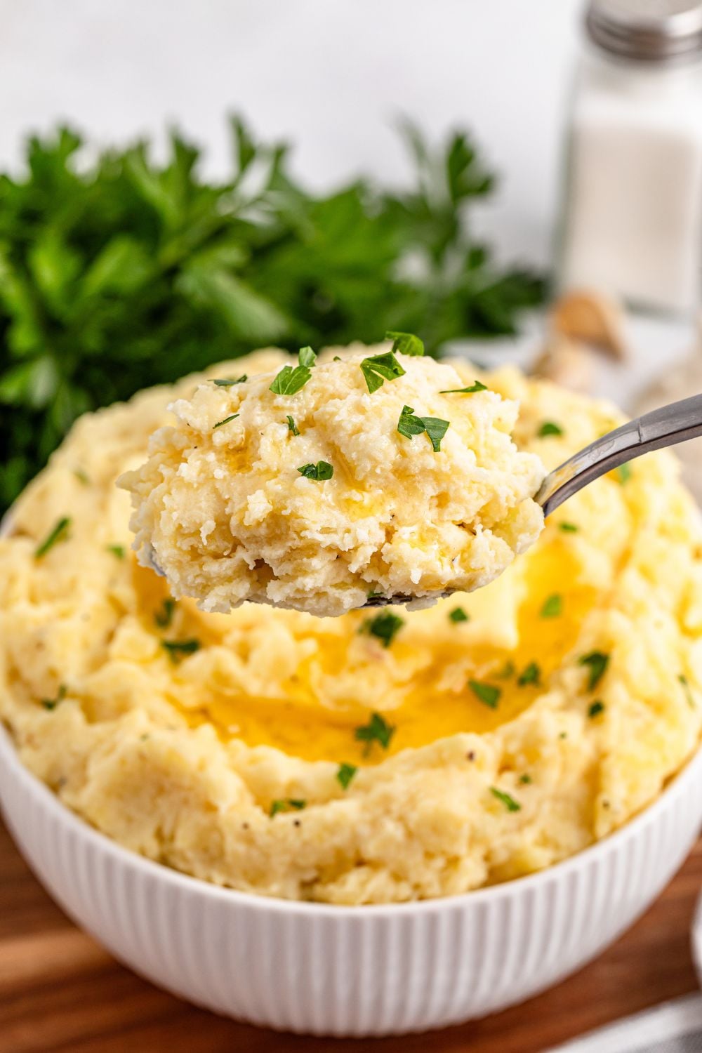 spoon full of Garlic Mashed Potatoes and a white bowl full of more potatoes behind it on a wooden cutting board sitting on a white table 