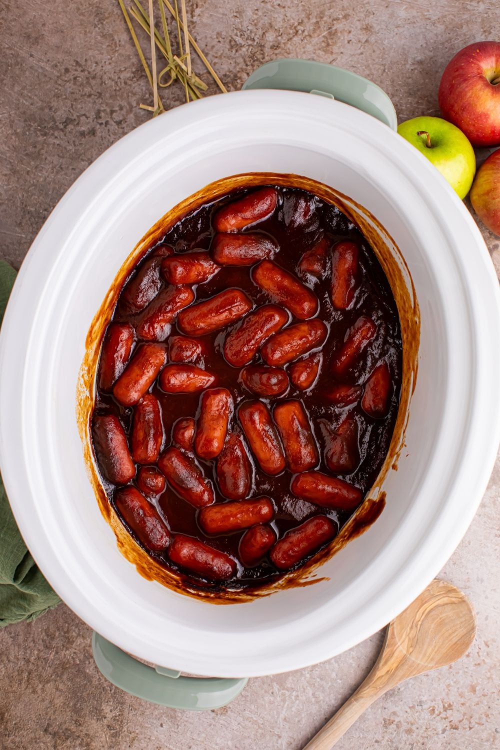 looking down on table with a white Crockpot full of Apple Butter BBQ Little Smokies with apples to the side of table and wooden spoon