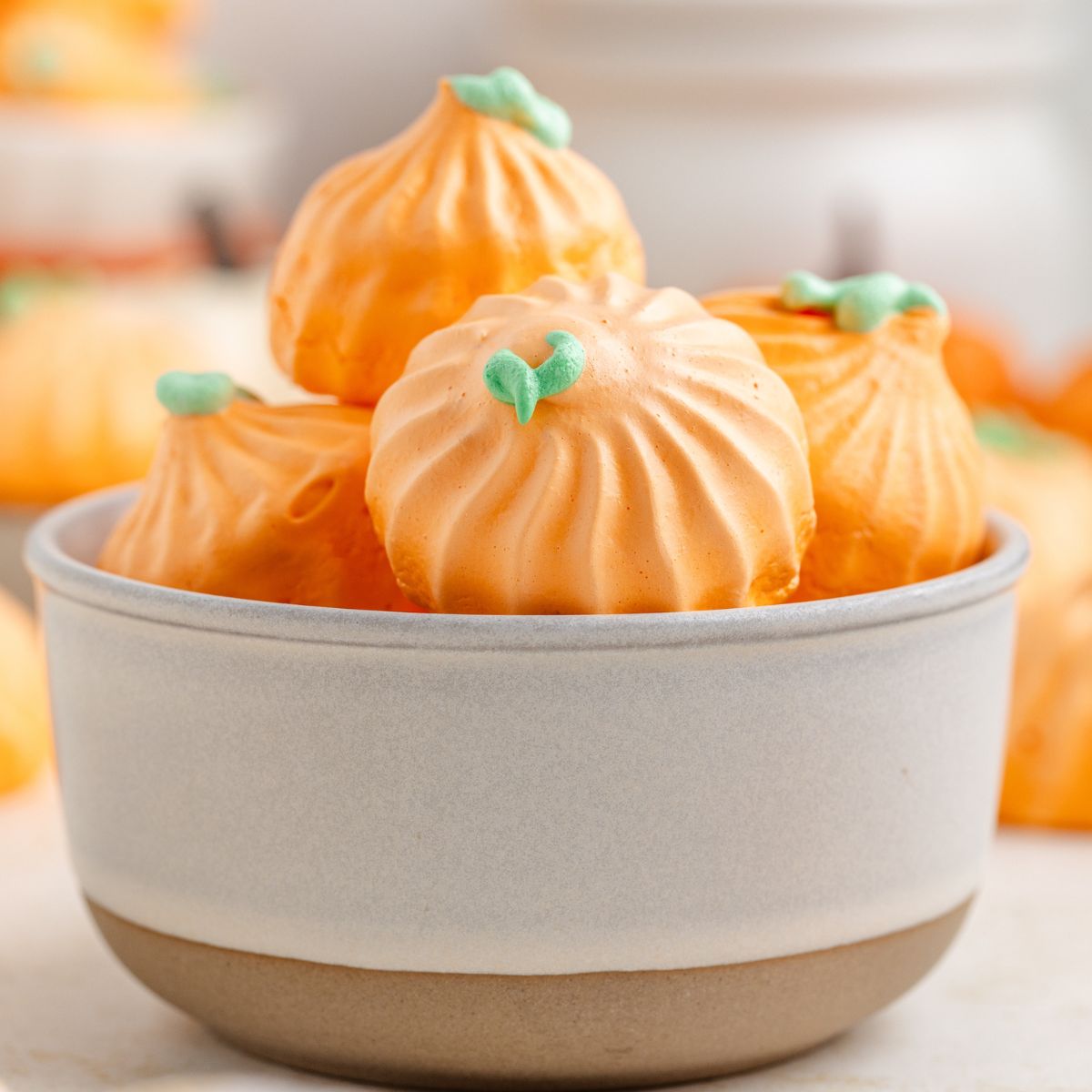 Pumpkin Meringues placed in a white bowl on table with more meringues behind it on table 