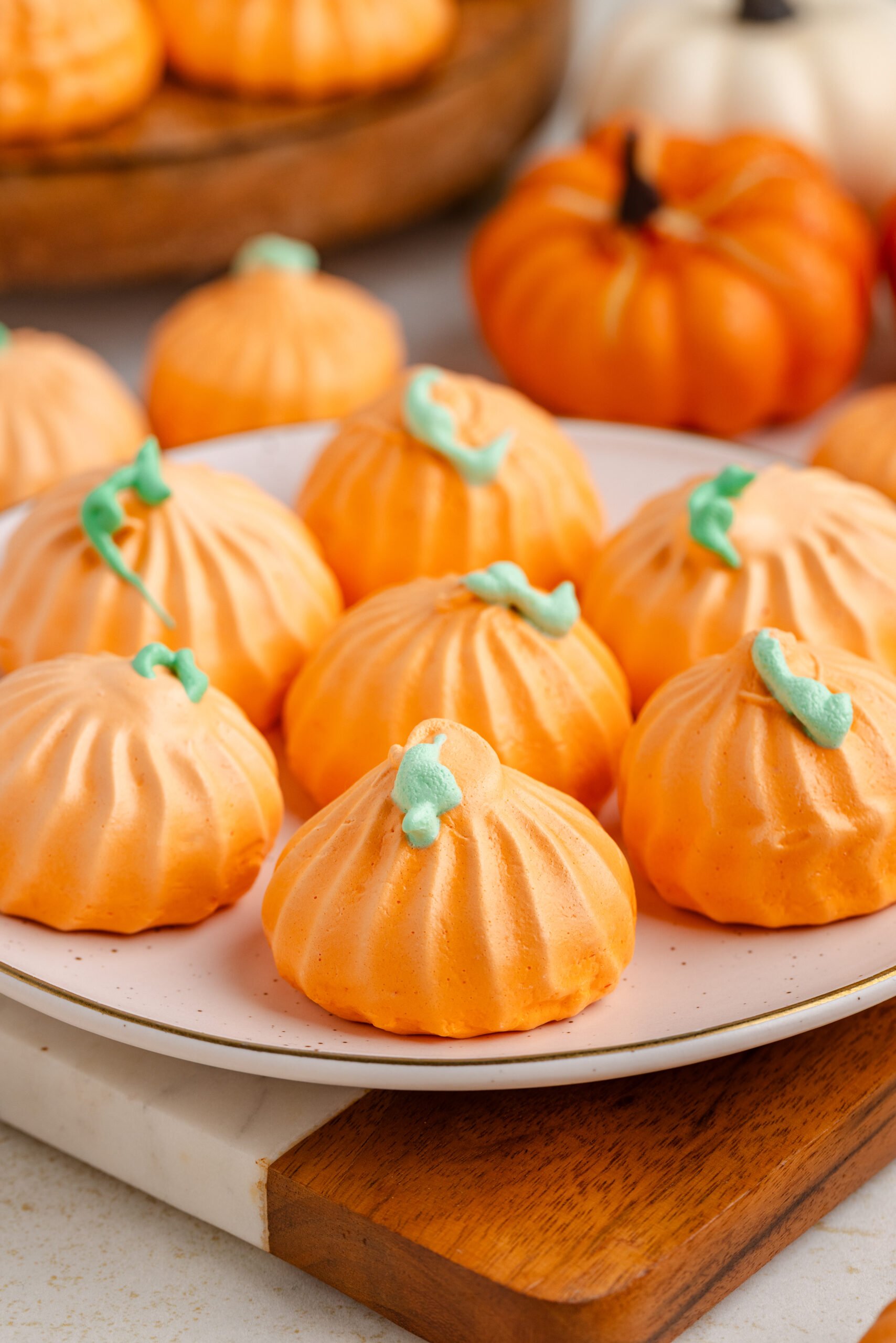 Pumpkin Meringue cookies on a plate with faux pumpkins behind it on table and a tray with more cookies 