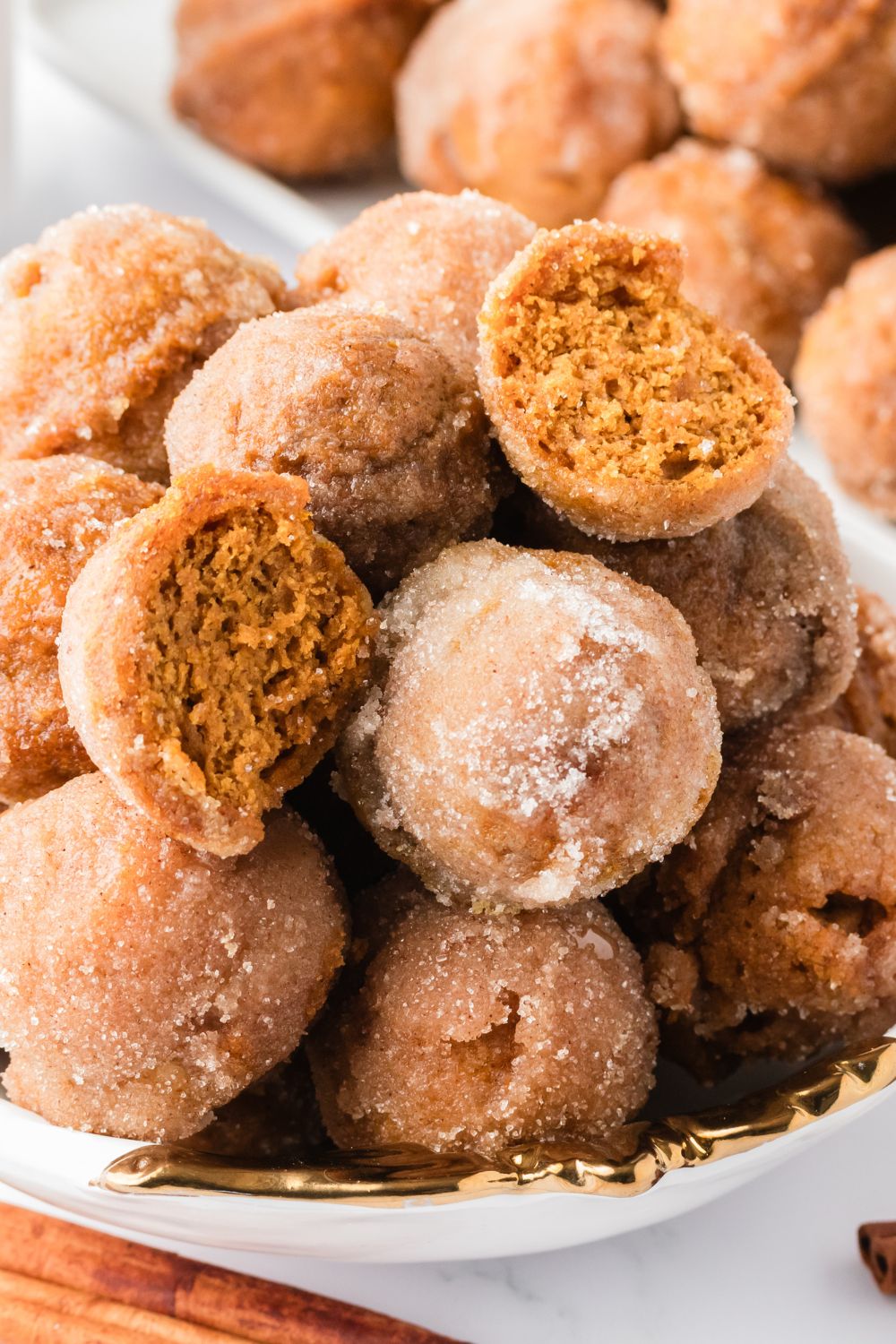 Pumpkin Spice Donut Holes stacked in a bowl on table with one cut in half, and a tray full of donuts behind it on table 