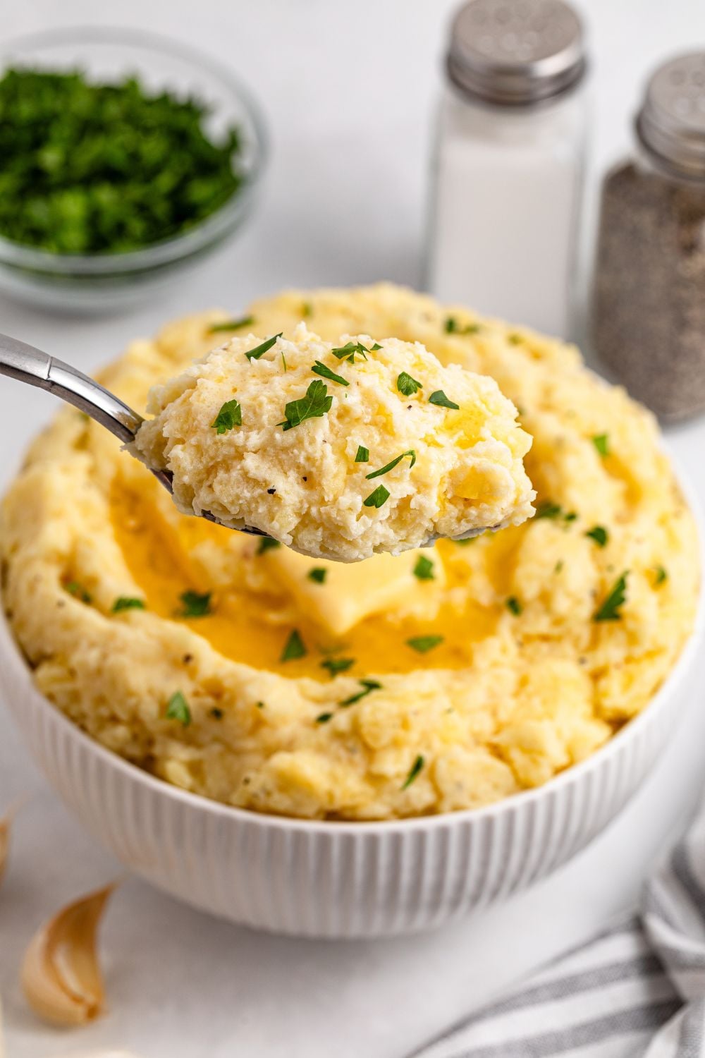 spoon full of Garlic Mashed Potatoes and a bowl behind it sitting on the white table with more potatoes. To the side is a salt and pepper shaker and small glass bowl with chopped parsley