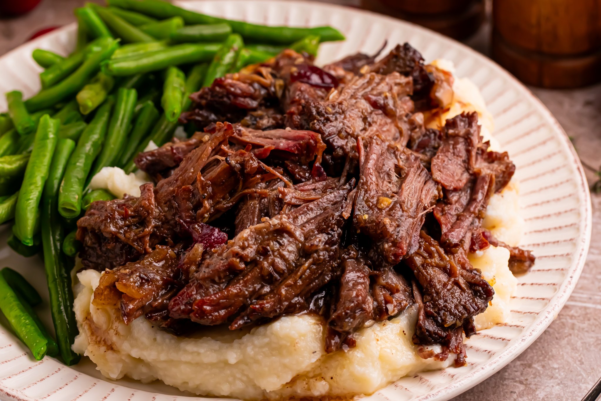 Juicy Cranberry Pot Roast shredded on a white plate with mashed potatoes under and a side of green beans 