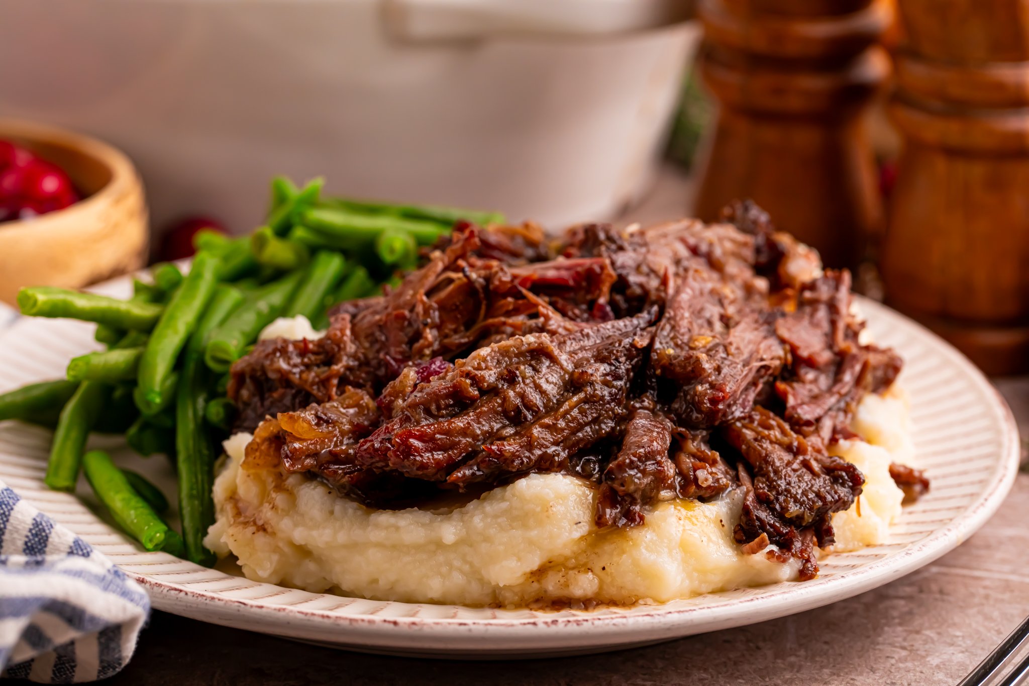 Side view of the Cranberry Pot Roast shredded on a white plate with mashed potatoes under and a side of green beans 