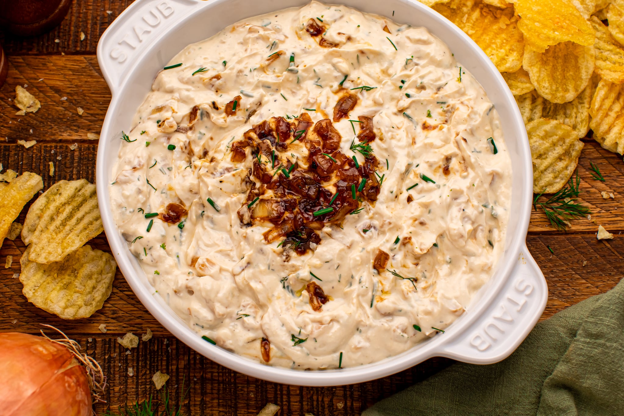Overhead photo of Caramelized Onion Chip Dip in a white staub bowl on dark wooden table with chips  all around the bowl