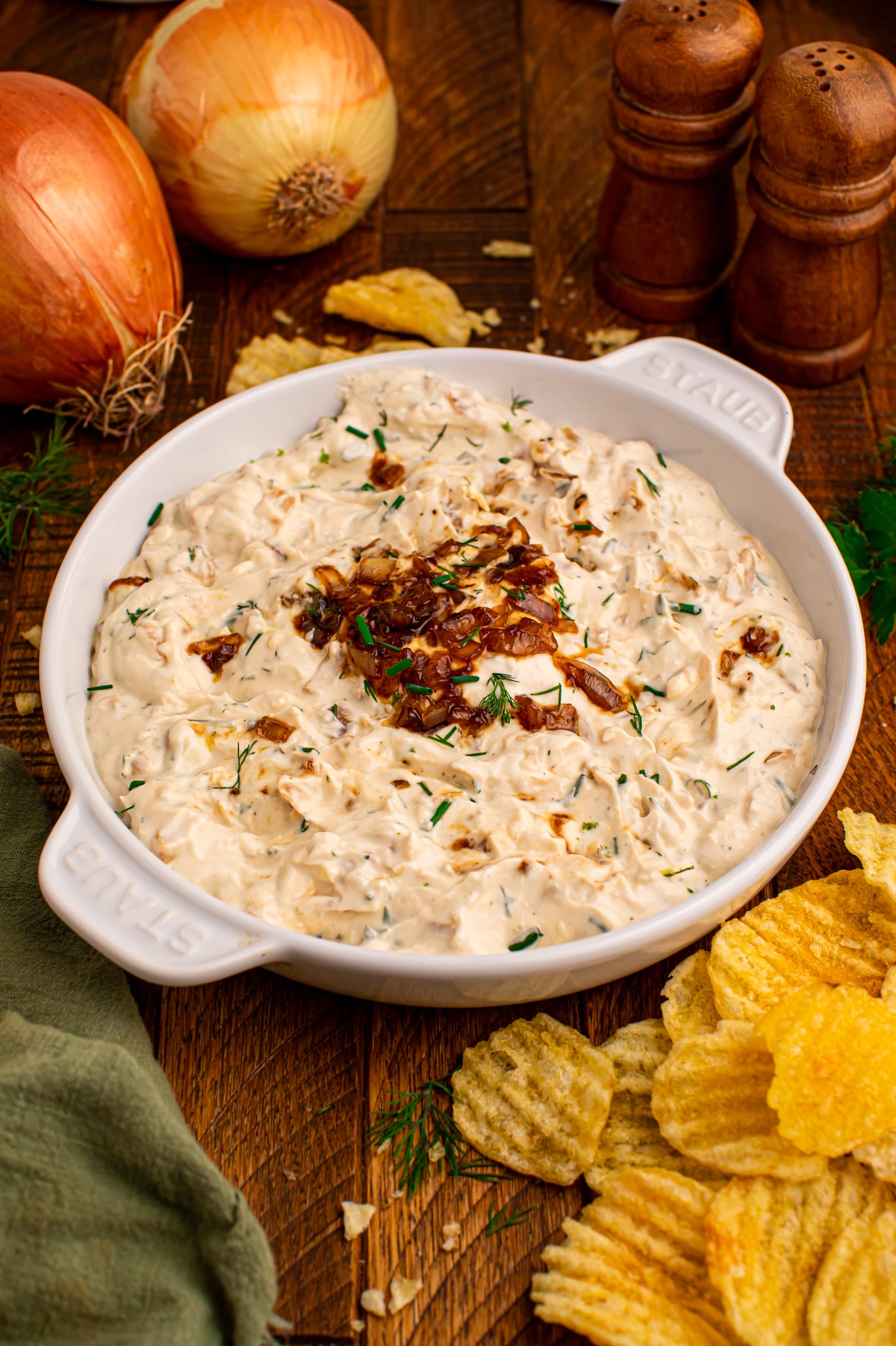 side view of the Caramelized Onion Chip Dip in a white staub bowl on dark wooden table with chips to the side 