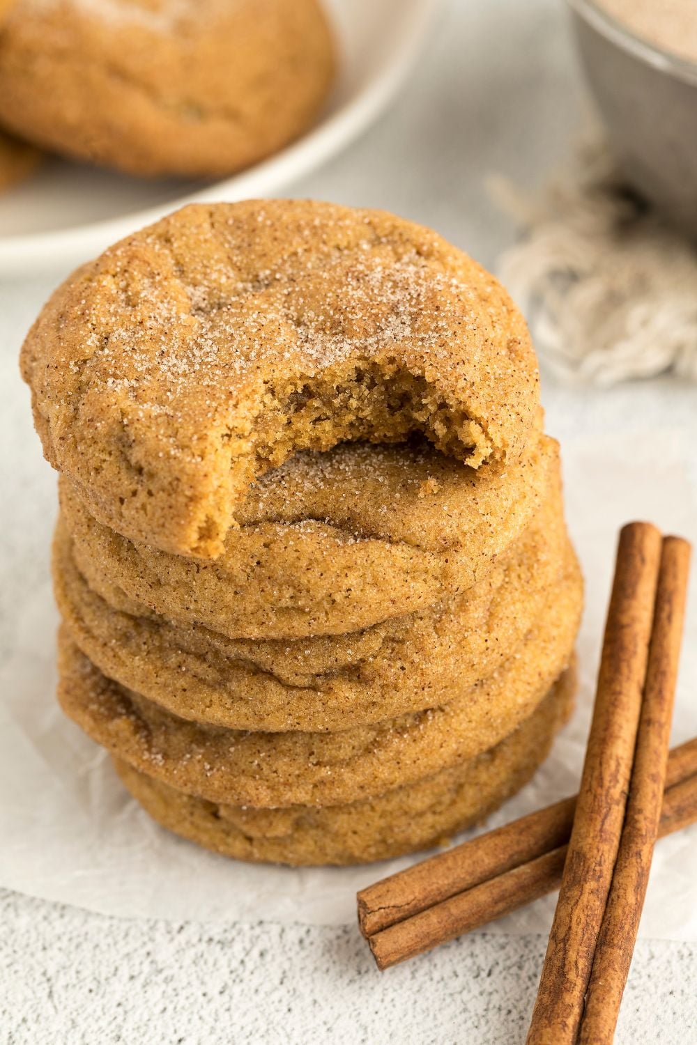 Pumpkin Snickerdoodle Cookies stacked on table with the top one with a bite taken out of it, and cinnamon sticks in front of cookies