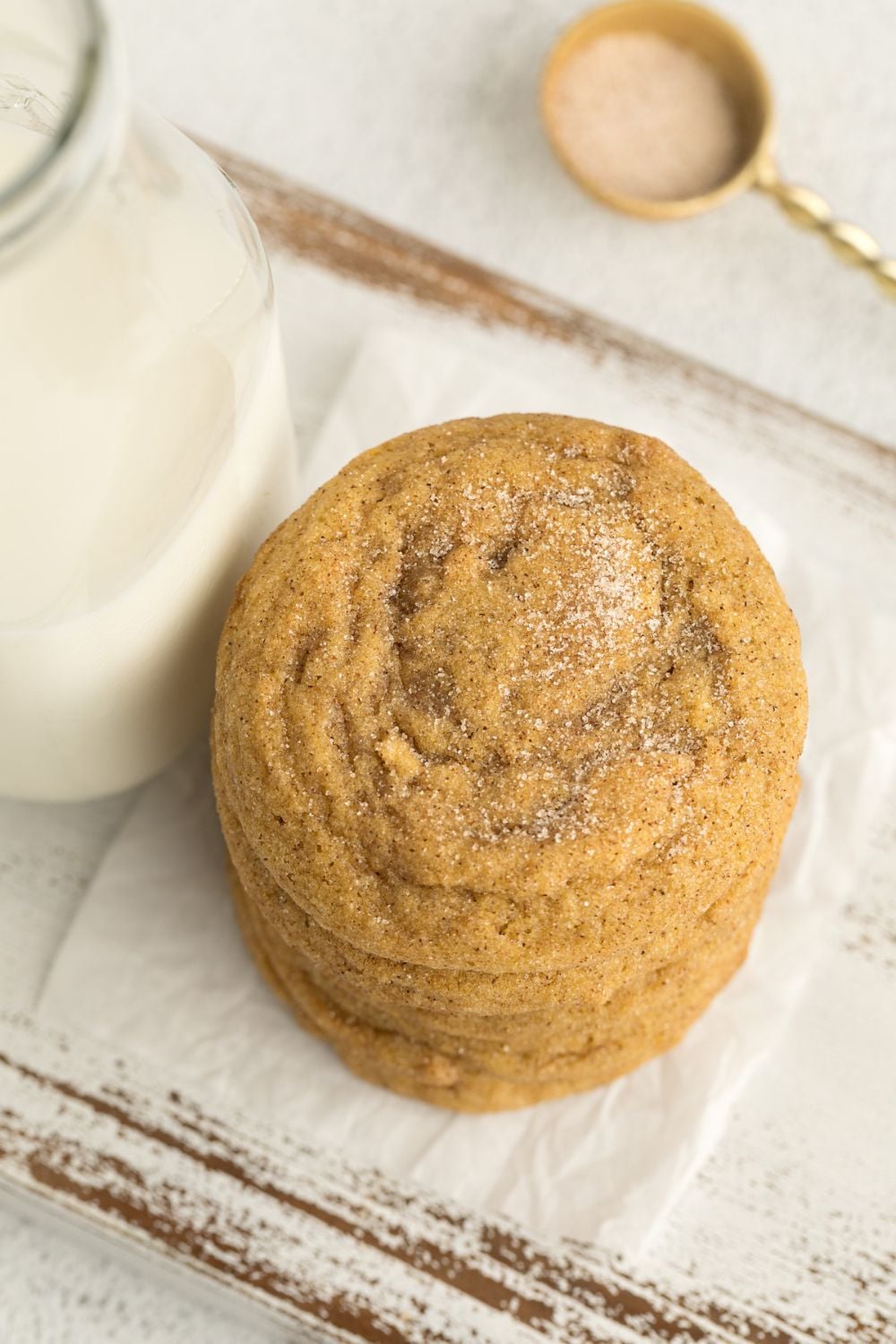 Pumpkin Snickerdoodle Cookies stacked on top of one another by a glass of milk and a small spoon of cinnamon sugar sitting behind it on table 