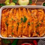 Steak Enchiladas in a casserole pan sitting on a wooden table with sliced limes, onion and cilantro behind the casserole dish
