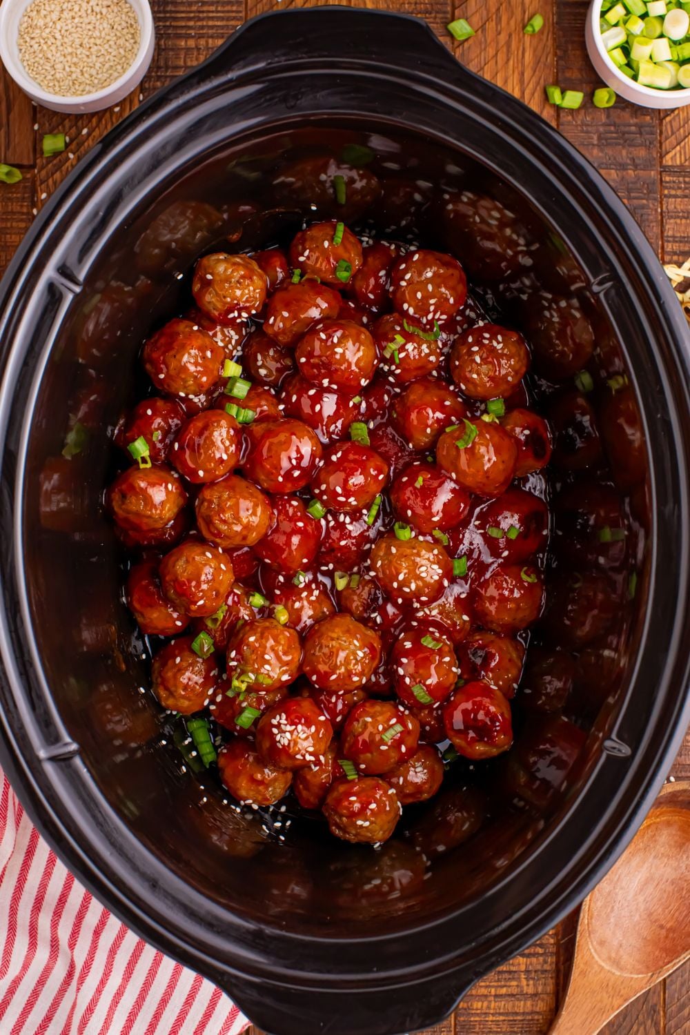 Crockpot Sweet Chili Meatballs in a black crockpot on wooden table with a red striped towel to side and a bowl of green onions 