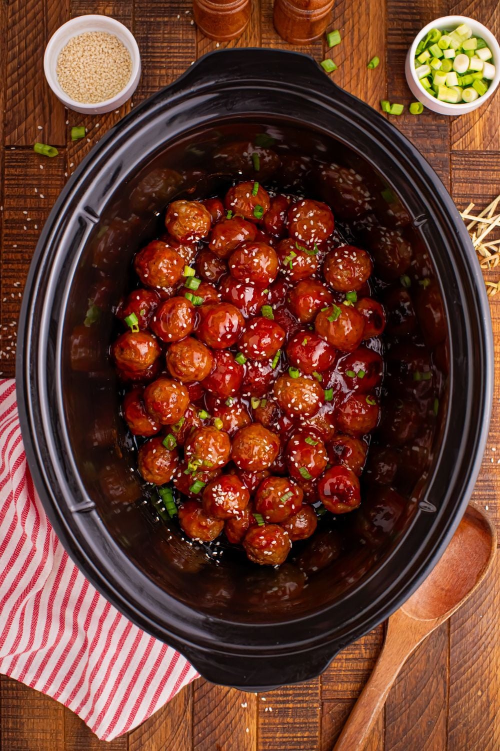 Crockpot Sweet Chili Meatballs on a wooden table with a wooden spoon and towel to side and in back bowls of sesame seeds and sliced green onions 