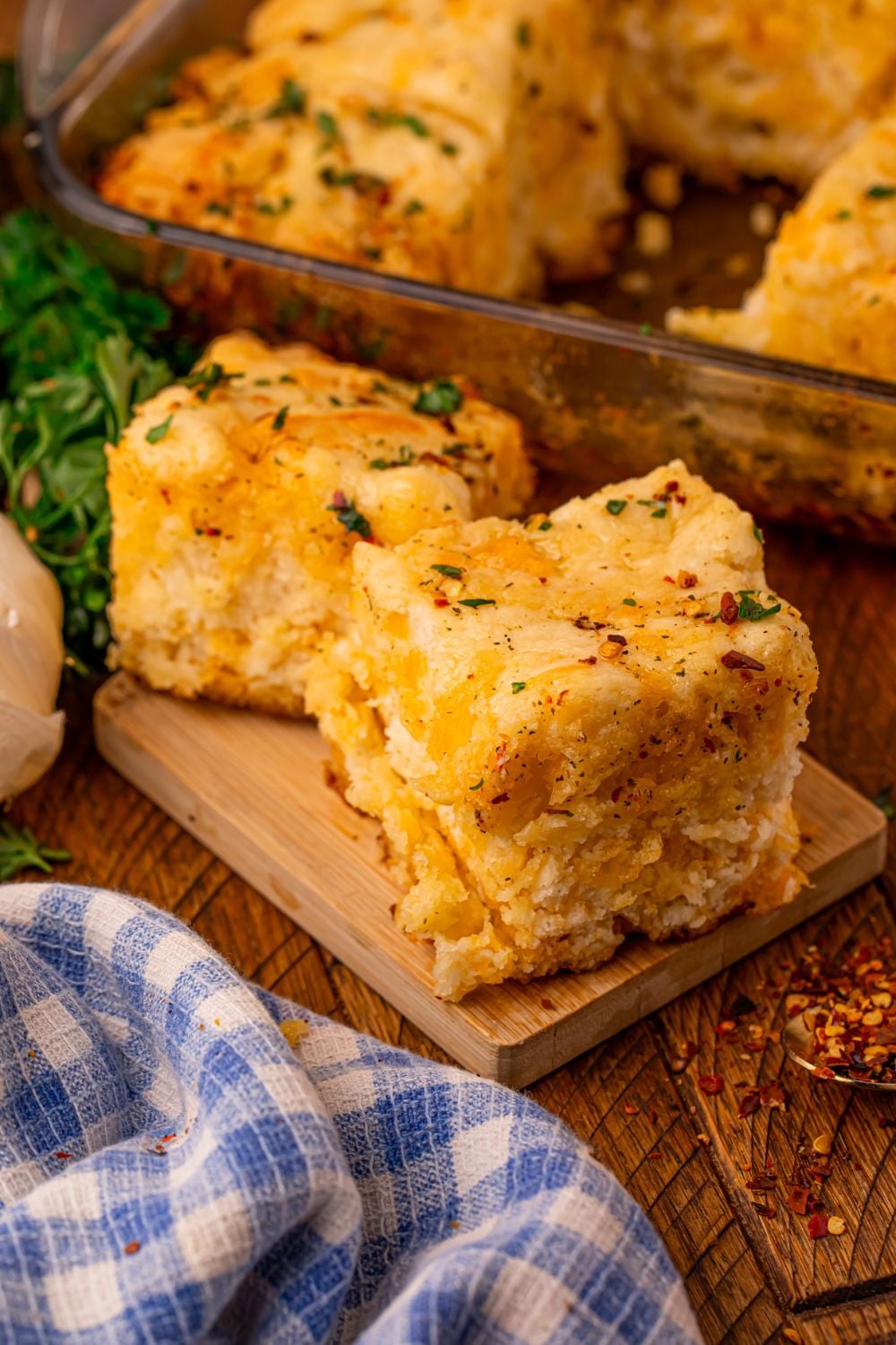Cowboy Butter Swim Biscuits with a glass pan with more biscuits behind the table, and a few placed in front of the table with a blue checkered towel in front 