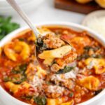 Sausage Tortellini Soup in a white bowl on table with a spoonful of soup just above the bowl