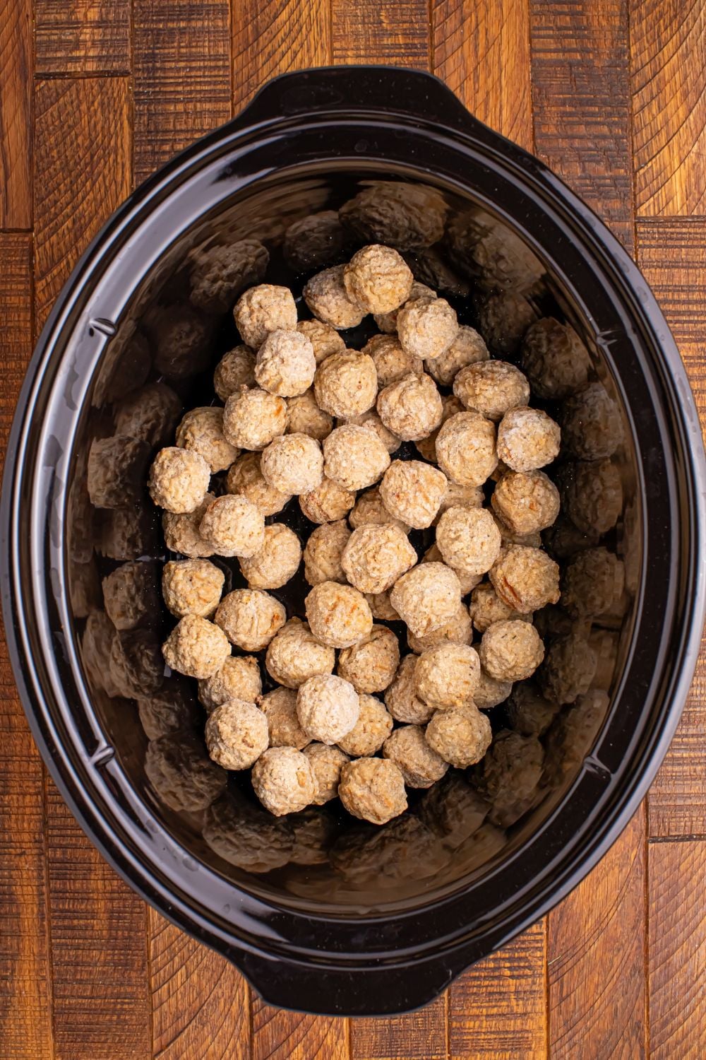 frozen meatballs in a black crockpot sitting on a wooden table for making Crockpot Sweet Chili Meatballs