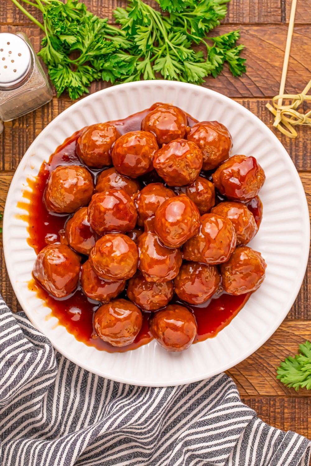 a wooden table with a white plate full of peach bbq meatballs, and salt and pepper to side, parsley, and toothpicks for serving up 