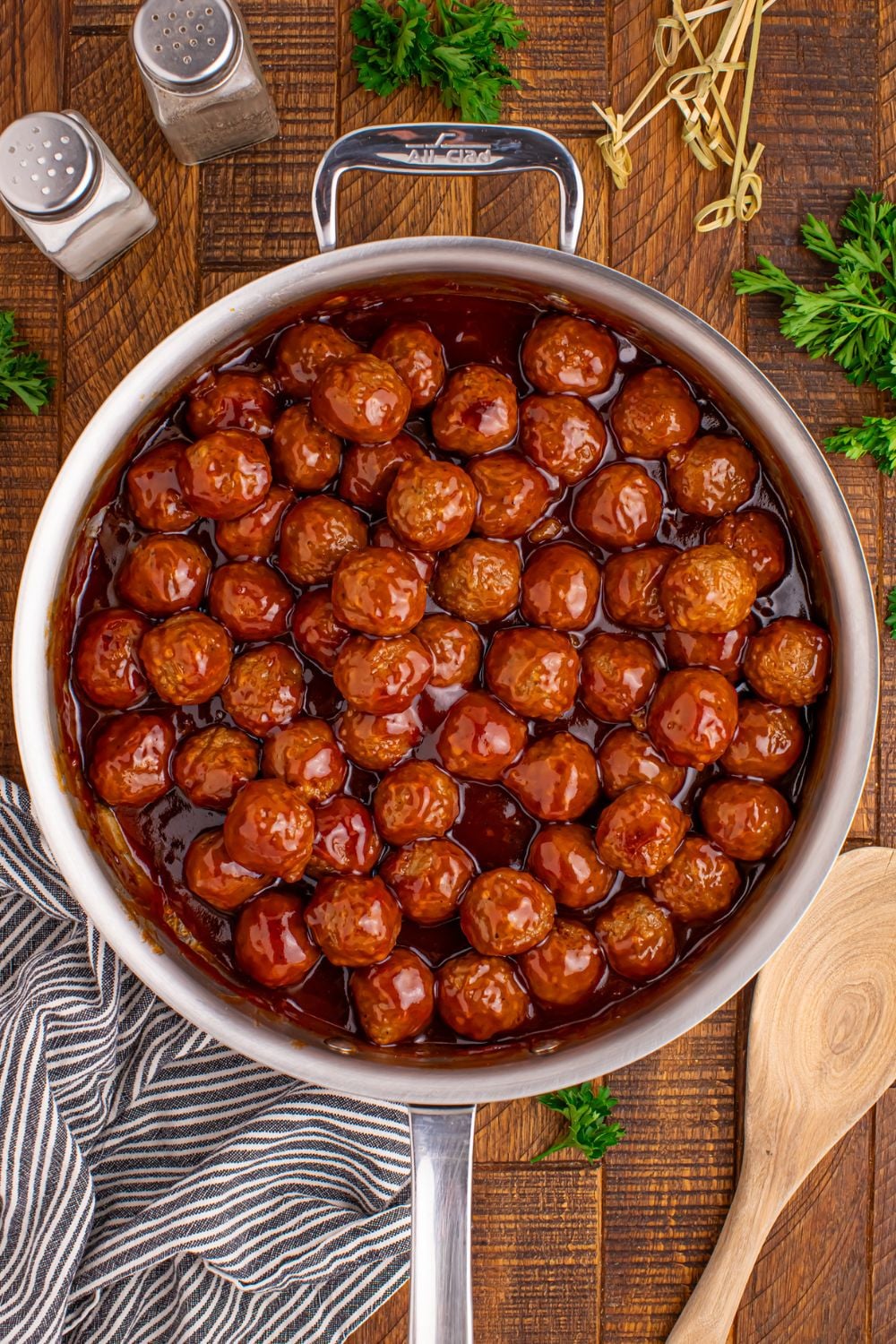 a large skillet on a wooden table full of cooked Peach BBQ Meatballs with salt and pepper shaker to side, parsley, and toothpicks scattered on table 