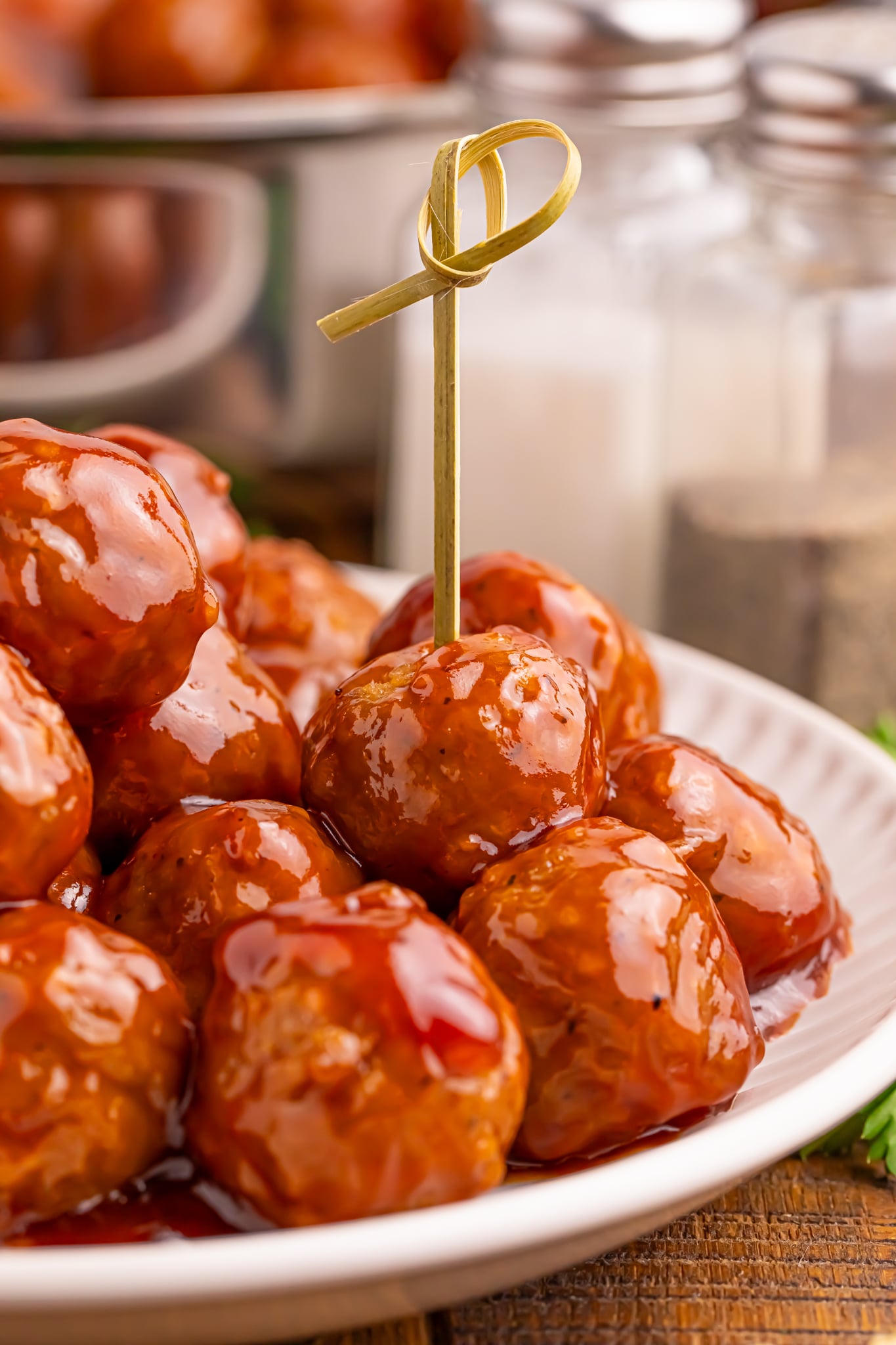 a white plate full of bbq peach meatballs with one that has a toothpick in it, and a salt and pepper shaker behind it on table