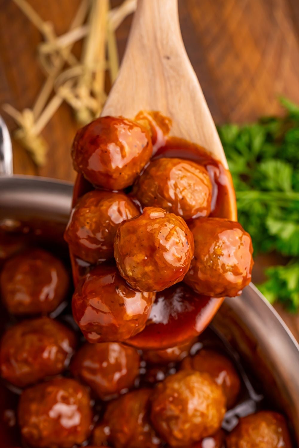 Peach BBQ Meatballs in a skillet with a wooden spoon full of meatballs and a bunch of parsley to side of skillet on wooden table