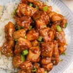 Bourbon Chicken piled on top of a plate with some rice on the side and topped with sesame seeds and green onions