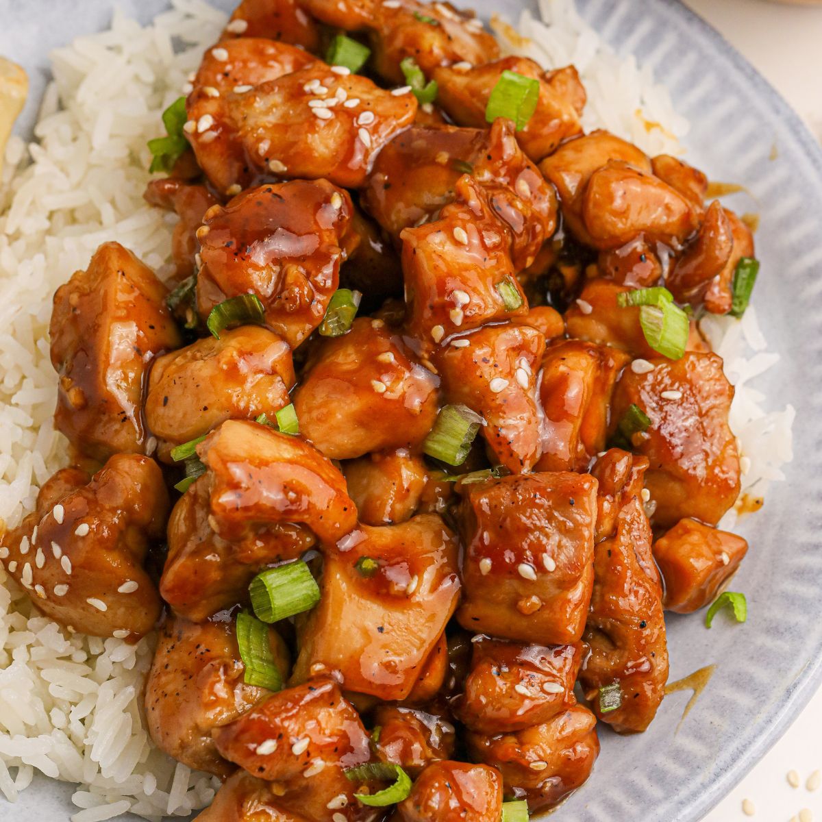 Bourbon Chicken piled on top of a plate with some rice on the side and topped with sesame seeds and green onions 